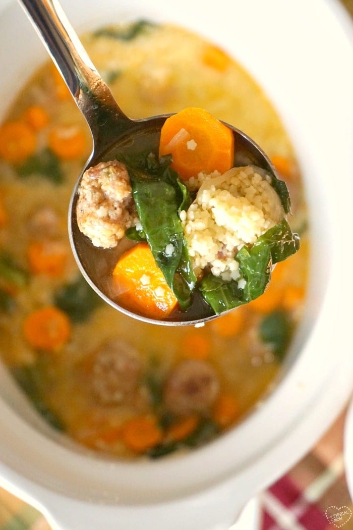 Close-up of a ladle holding a comforting slow cooker soup with carrots, kale, couscous, and meatballs over a white pot.