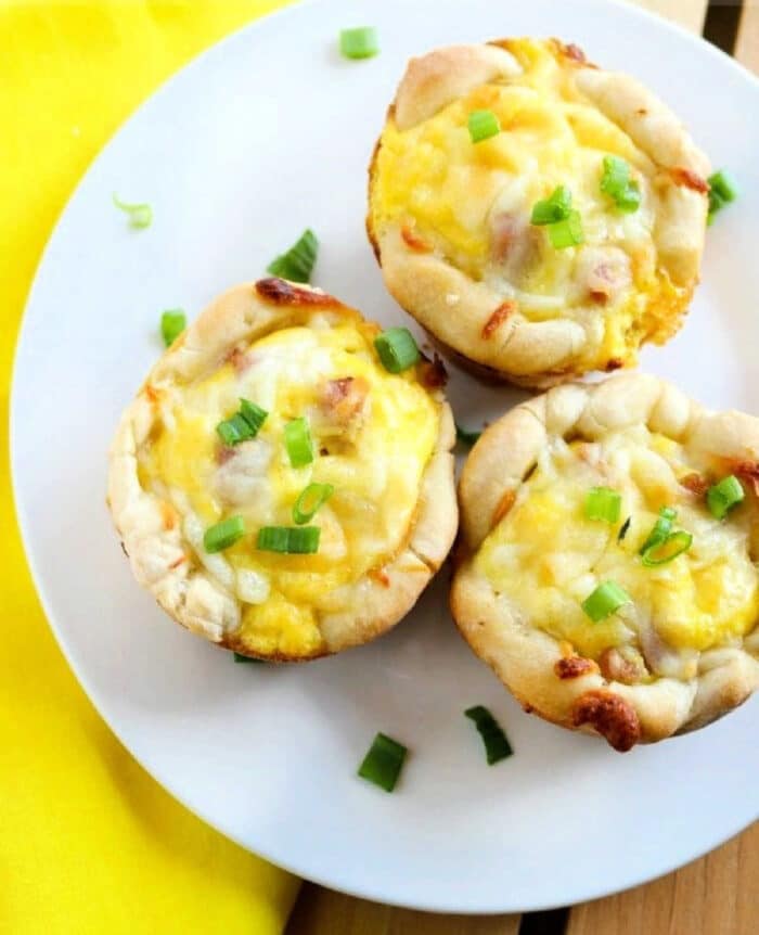 Three baked breakfast muffins filled with cheese, eggs, and green onions are arranged on a white plate. They sit beside a yellow napkin, reminiscent of delicious ham and cheese biscuits.