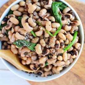 A wooden spoon rests on a bowl of seasoned black-eyed peas with spinach, inviting you to savor its flavor. Wondering how long to cook black-eyed peas? Perfectly tender in just about an hour, they make a delicious and wholesome meal on any wooden surface.