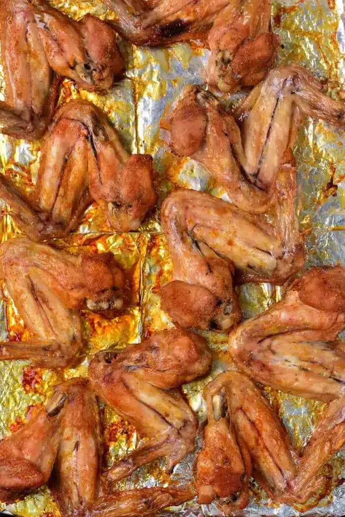 A baking sheet is covered with a layer of baked whole chicken wings. The wings are arranged in rows on a foil-lined tray and appear to be seasoned and baked until golden brown.