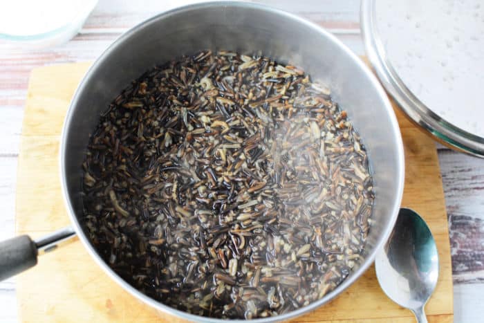 A pot of wild rice cooking on a wooden cutting board, steam rising invitingly. Next to it, a stainless steel bowl and a silver spoon await. In the background, the white surface adds a crisp contrast. Ever wondered how long does rice take to cook? This pot holds the answer in its fragrant steam.