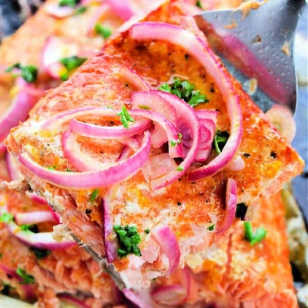 A close-up of a slice of grilled salmon topped with sliced red onions and chopped green herbs. The piece, baked to perfection at 400°F, is being lifted with a metal spatula. More pieces of grilled salmon are visible in the background, showcasing the delicious outcome.