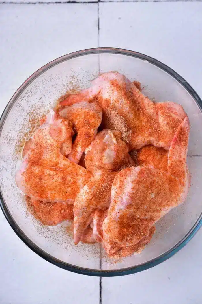 A glass bowl containing raw chicken wings, seasoned with a reddish spice mix, ready to be transformed into delicious baked whole chicken wings, sits on a white tiled surface.