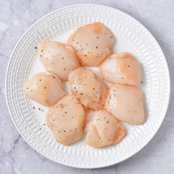 A white plate with raw, seasoned scallops rests on a marble surface, inviting curiosity about how long to cook these delicate treasures to perfection.