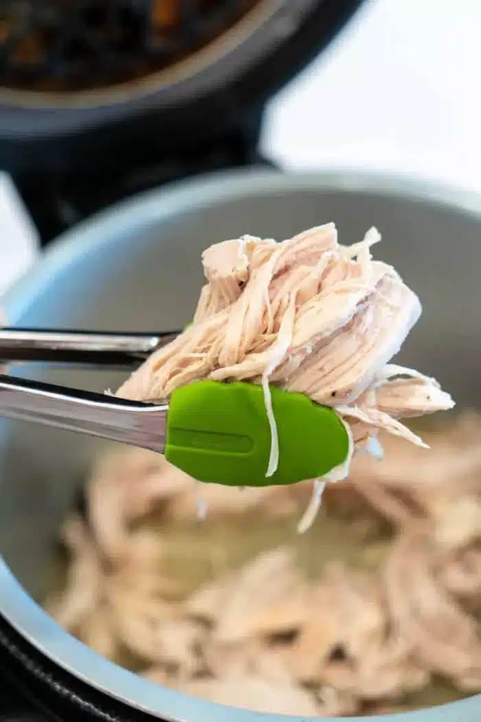 Tongs with green silicone tips hold Ninja Foodi shredded chicken over an open pressure cooker. The light-colored chicken appears freshly cooked, while the background reveals more juicy, tender goodness inside the cooker.