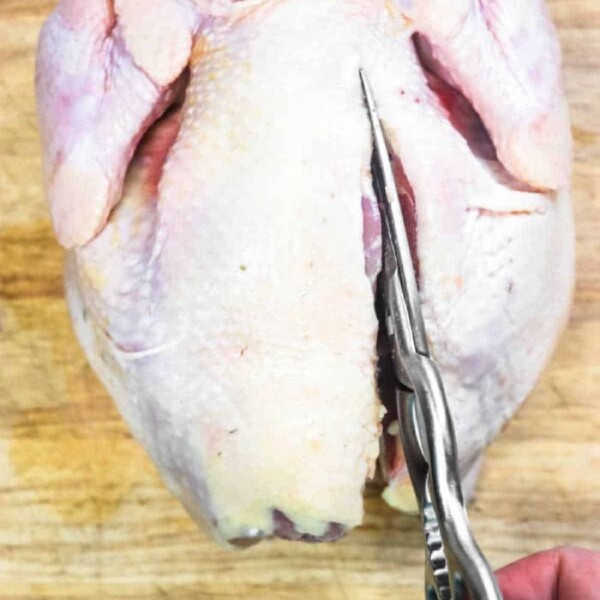 A pair of kitchen shears is cutting through the back of a whole raw chicken placed on a wooden cutting board, demonstrating what is a spatchcocked chicken.