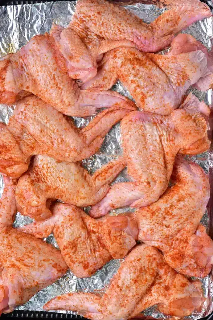 Raw, seasoned whole chicken wings arranged on a foil-lined baking sheet, ready to be baked. The wings are covered with a red spice rub.