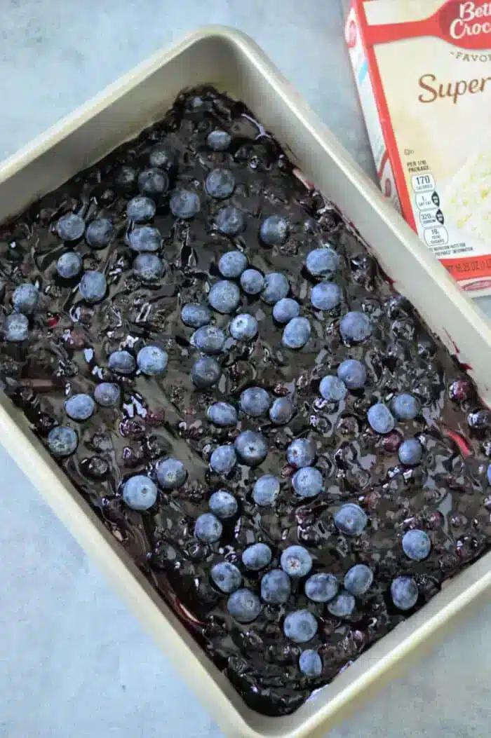 A baking pan brimming with blueberry pie filling, topped with fresh blueberries, showcases a delectable blueberry cobbler made with Betty Crocker Super Moist white cake mix. The partially visible box hints at the delightful blend that elevates this dessert on the gray surface.