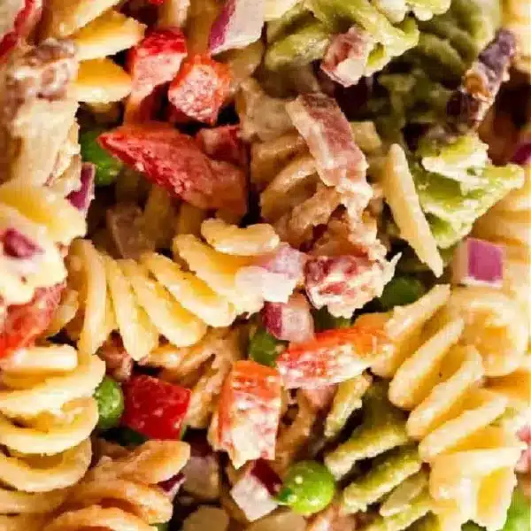 Close-up image of a vibrant bacon pasta salad featuring tri-color rotini, chopped red bell peppers, purple onions, peas, and crispy bacon pieces, all covered in a creamy dressing.