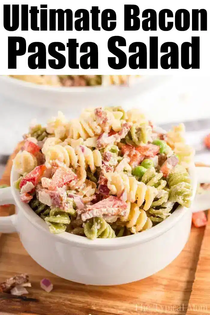 A white bowl filled with a colorful bacon pasta salad, which includes rotini, bacon, peas, and diced red bell peppers, is set on a wooden surface. The words "Ultimate Bacon Pasta Salad" are displayed at the top in bold black letters.