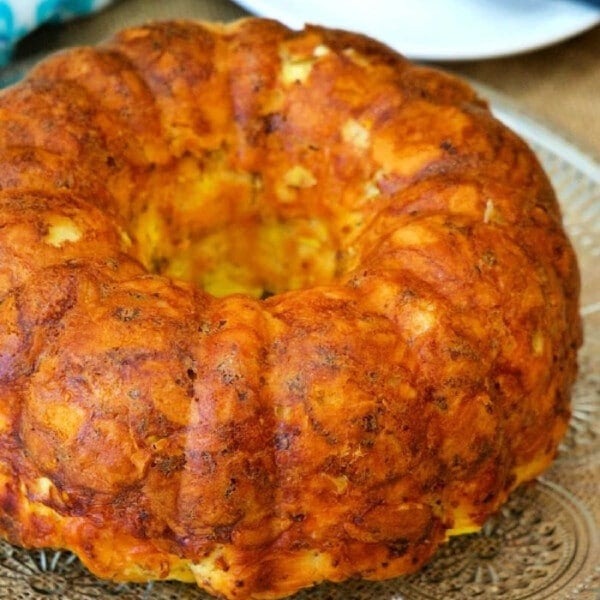 A golden brown breakfast bundt cake sits elegantly on a glass platter, with a knife poised nearby and a blue napkin adding a touch of color in the background.