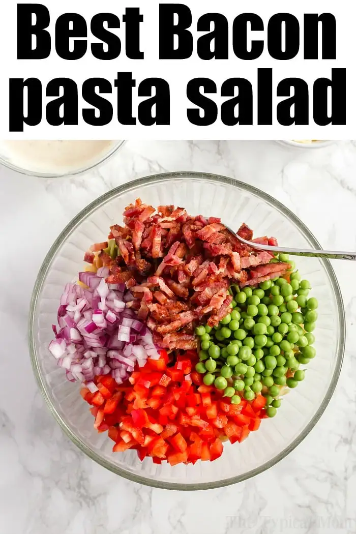 A glass bowl filled with chopped ingredients for a bacon pasta salad: crispy bacon, red onions, peas, and red bell peppers. Above the bowl, text reads "Best Bacon Pasta Salad." The background is a pristine white marble surface.