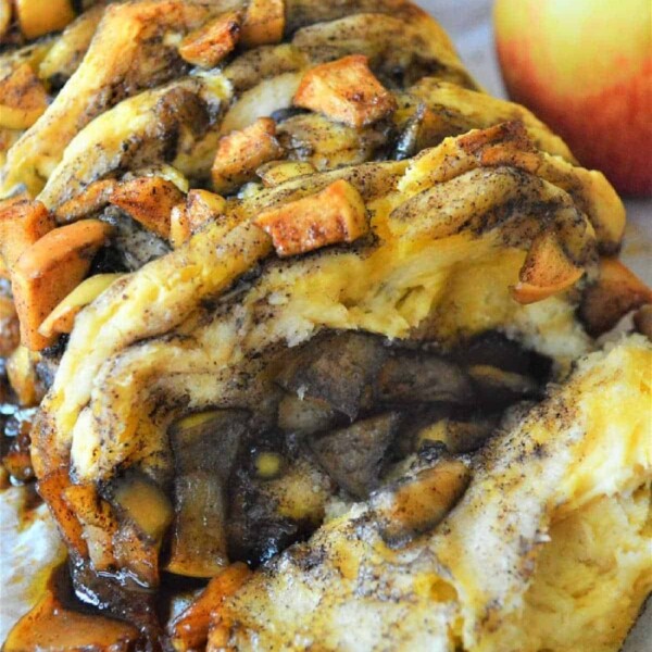 Close-up of a sliced cinnamon pull apart bread on parchment paper, topped with chunks of apples and swirls of cinnamon. An apple sits in the background.