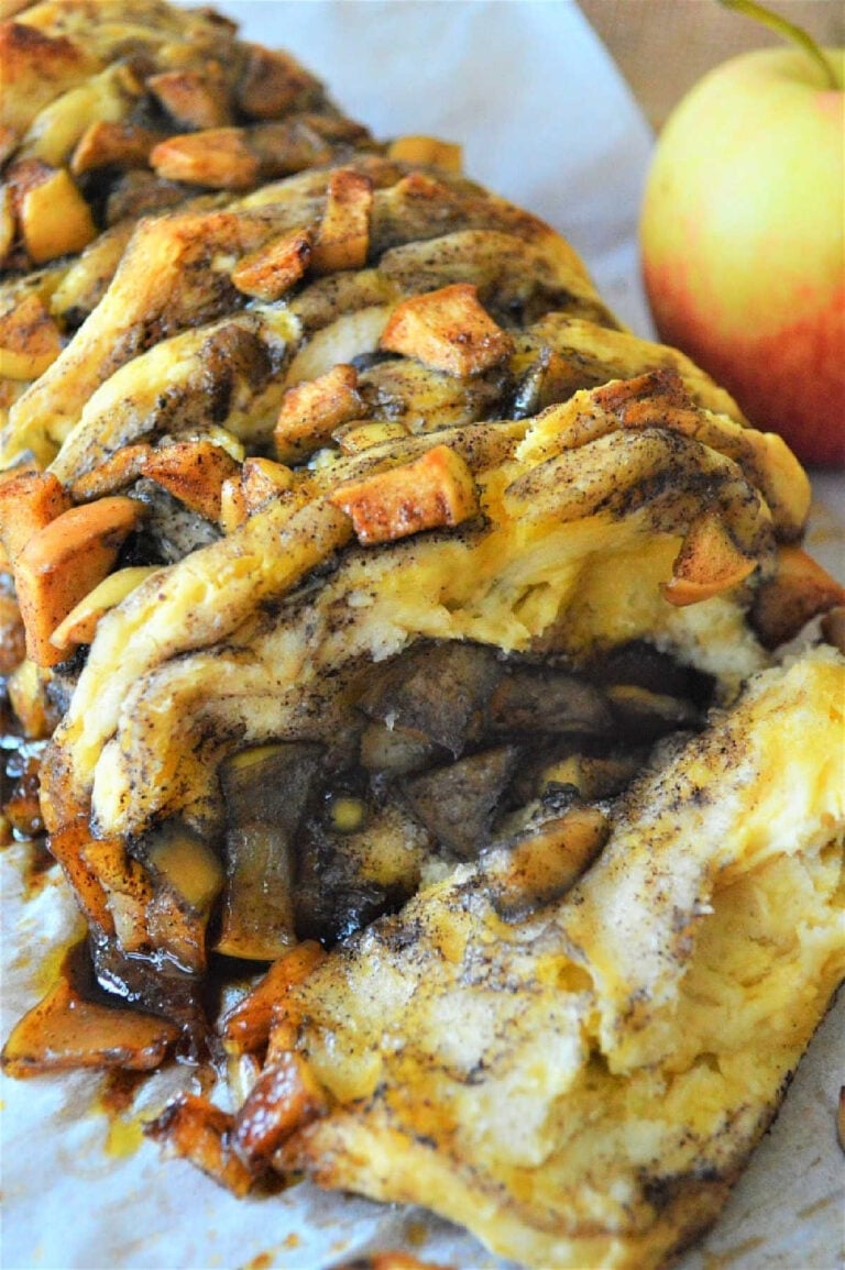 Close-up of a sliced cinnamon pull apart bread on parchment paper, topped with chunks of apples and swirls of cinnamon. An apple sits in the background.