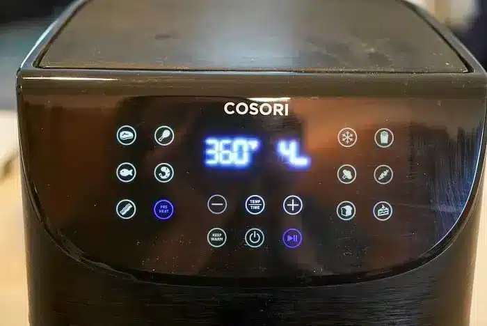 An air fryer with a digital display set to 360 degrees Fahrenheit and 4 minutes. Various icons surround the display indicating different cooking modes and functions, guided by an air fryer cooking times chart. The brand name "COSORI" is visible above the controls.