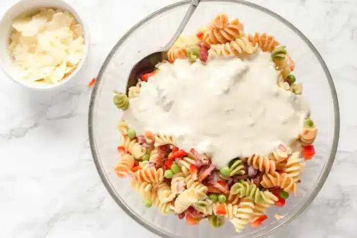 A clear glass bowl filled with colorful rotini pasta, peas, diced red bell peppers, and cubes of ham forms the heart of this enticing bacon pasta salad. A white creamy sauce is poured on top. A metal spoon is in the bowl. A small bowl beside it contains shaved Parmesan cheese.