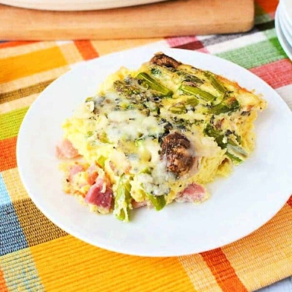 A slice of vegetable asparagus egg casserole recipe, rests on a white plate atop a colorful plaid tablecloth.