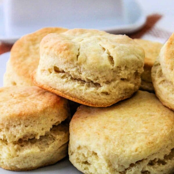 A plate of freshly baked air fryer biscuits stacked on top of each other. The golden brown biscuits boast a soft, fluffy texture. A butter dish is blurred in the background.