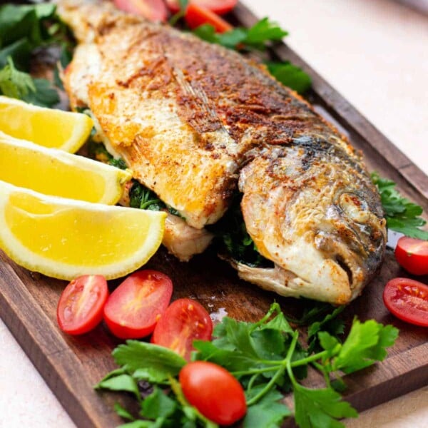 A cooked whole fish is served on a wooden board, garnished with lemon wedges, cherry tomatoes, and parsley. The fish has a seasoned, crispy golden-brown skin. Fresh parsley and halved cherry tomatoes are placed around the fish for decoration; it's an inviting way to fry a whole fish to perfection.