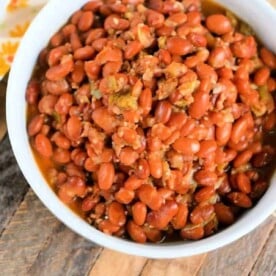 A white bowl filled with seasoned pinto beans, fresh from the pressure cooker, sits on a wooden surface. The reddish-brown beans are tantalizingly presented alongside a sliver of floral-patterned cloth peeking in from the top left corner.