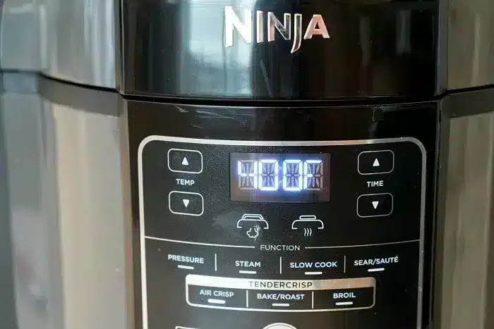 Close-up of a Ninja multi-function kitchen appliance. The digital display shows "4:00." Buttons for various functions including Temp, Time, Pressure, Steam, Slow Cook, Sear/Sauté, Air Crisp, Bake/Roast, and Broil are visible below the display. Refer to the air fryer cooking times chart for precise results.