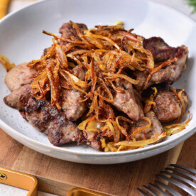 A bowl of expertly cooked liver, topped with crispy fried onions, illustrates how to cook organ meat to perfection. Presented on a rustic wooden cutting board, this dish promises a delectable experience for those eager to explore the culinary world of organ meats.