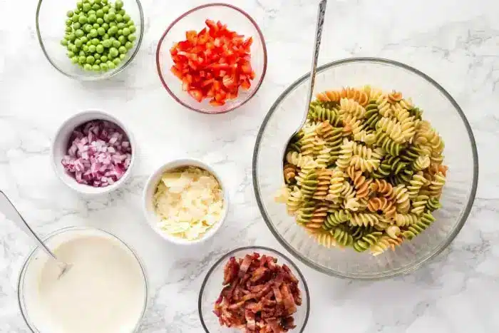 A variety of ingredients are laid out in small bowls on a white marble countertop. There is a large bowl of tri-color rotini pasta for the bacon pasta salad, peas, chopped red bell pepper, red onion, parmesan cheese, bacon pieces, and a bowl of creamy sauce. A spoon is in the pasta bowl.