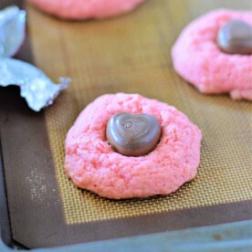 A delightful pink cookie, crafted from a strawberry cake mix, rests with a heart-shaped chocolate atop a baking sheet.