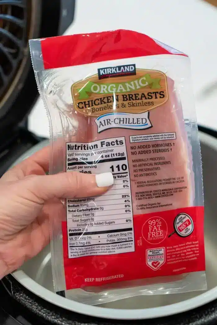 A package of Kirkland organic chicken breasts is held by a hand over a Ninja Foodi, ready to transform into delicious shredded chicken. The packaging, labeled as boneless, skinless, and air-chilled, displays nutritional facts and other details.