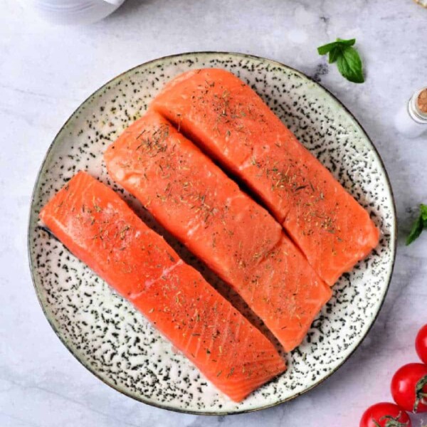 Three raw sockeye salmon fillets rest on a speckled plate with herbs. Nearby are fresh tomatoes, a bowl of garlic cloves, and a small pitcher of cream—perfect ingredients for those wondering how to cook sockeye salmon to luscious perfection.