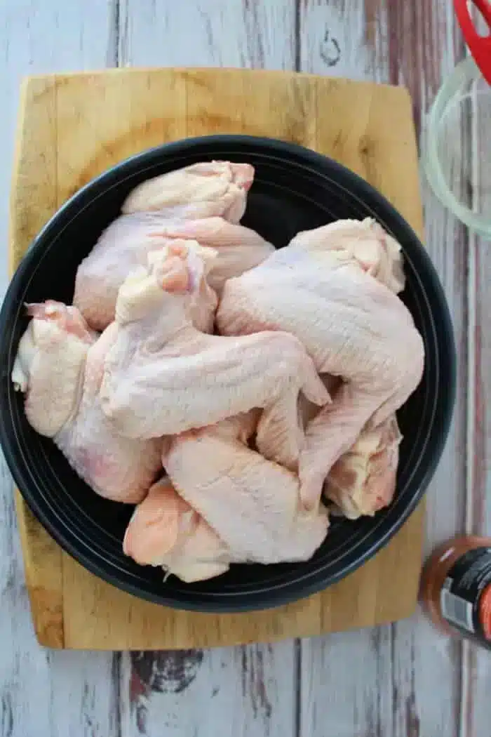 A top view of a black plate filled with raw chicken wings, ready to be transformed into delicious baked whole chicken wings, placed on a wooden chopping board. The background features a rustic wooden surface, a small glass, and part of a container of spices.