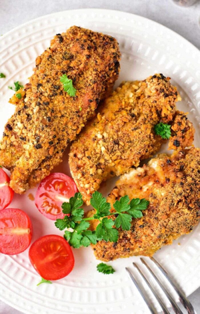 A plate with three almond-crusted, breaded, and baked chicken tenders garnished with parsley. Sliced cherry tomatoes are placed on the side. A fork is partially visible at the bottom right of the plate.