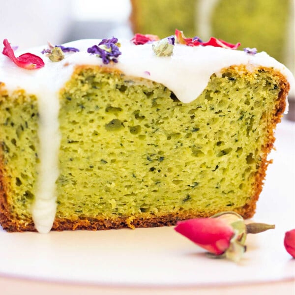 Close-up of a avocado cake slice with white icing and dried flower petals on top, complemented by rosebuds nearby, artfully reminiscent of an avocado cake.