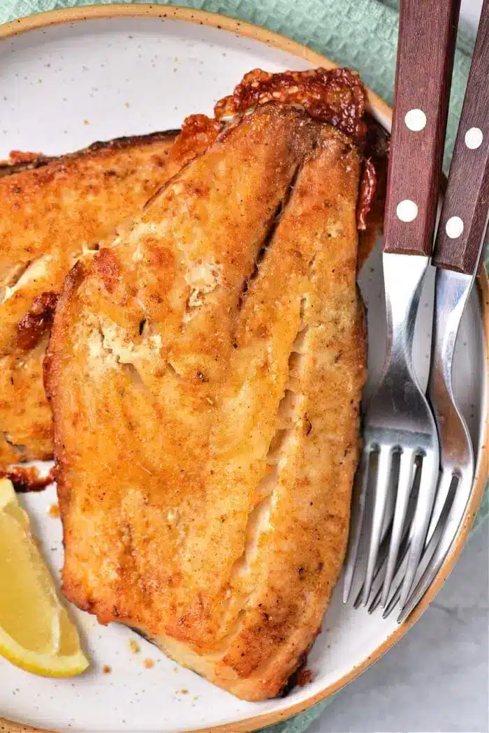 Grilled fish fillets with a golden crust rest on a white plate, alongside a lemon wedge. Perfectly cooked, just like baking frozen fish to perfection. A set of silverware with wooden handles is placed beside them, with a green textured cloth partially visible in the background.