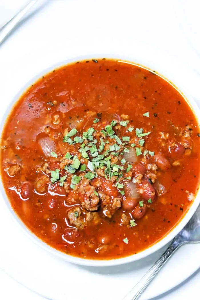 A bowl of chunky bear meat chili topped with chopped herbs. The hearty mix features beans, onions, and tender ground meat, all served in a pristine white bowl with a spoon ready on the side.