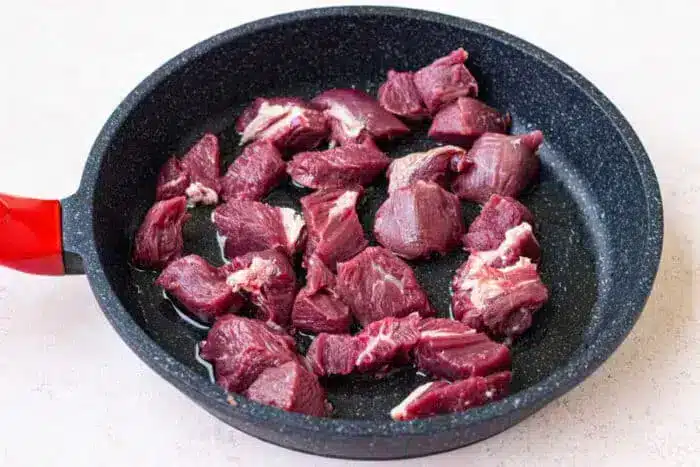 Raw chunks of bear meat are placed in a black frying pan with a red handle, resting on a light-colored countertop.