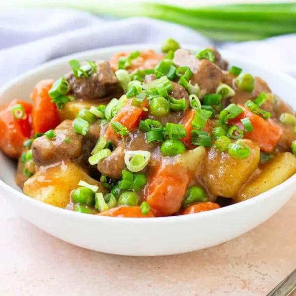 A bowl filled with a colorful stew containing chunks of bear meat, potatoes, carrots, and peas. The dish is garnished with chopped green onions. A silver fork lies next to the bowl, and a light-colored napkin is partially visible in the background.