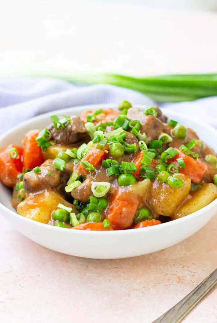 A white bowl filled with a colorful dish featuring tender bear meat chunks, carrots, potatoes, and peas, garnished with chopped green onions. A bunch of whole green onions is in the background.