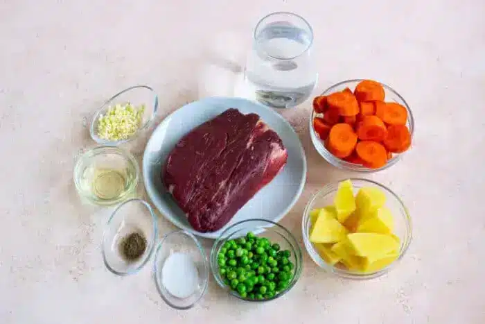 A raw piece of beef, reminiscent of bear meat in its robust appearance, rests on a plate surrounded by bowls of diced carrots, chopped potatoes, green peas, minced garlic, salt, pepper, oil, and a glass of water on a beige surface.