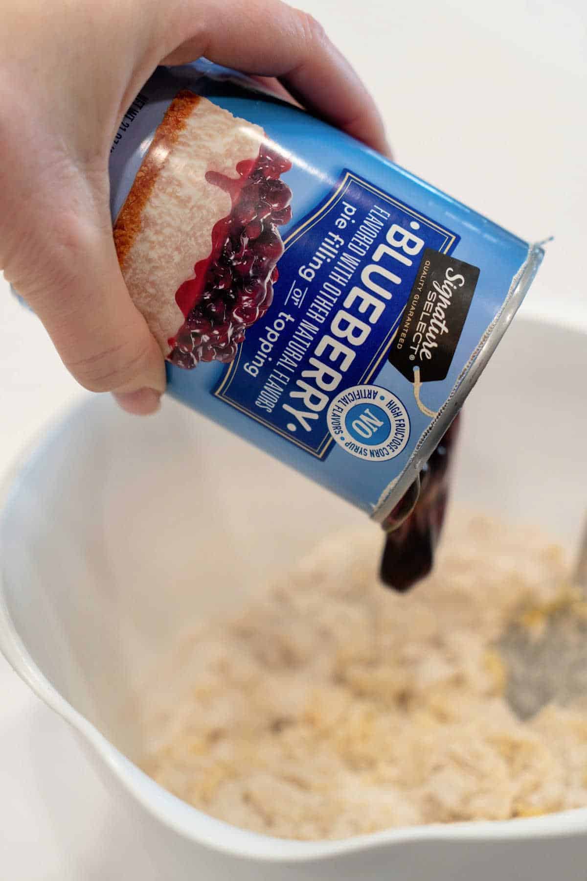 A hand is pouring blueberry canned pie filling into a white mixing bowl containing dry ingredients, perfect for any blueberry canned pie filling recipes. The can’s label reads “Blueberry,” and it displays an image of cheesecake with blueberry topping. The bowl rests on a light-colored surface.