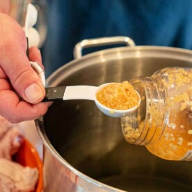 A person holds a jar of minced garlic, using a white measuring spoon to scoop it into a large metal pot for their whole chicken brine. A plastic bag with uncooked chicken wings is visible in the background.