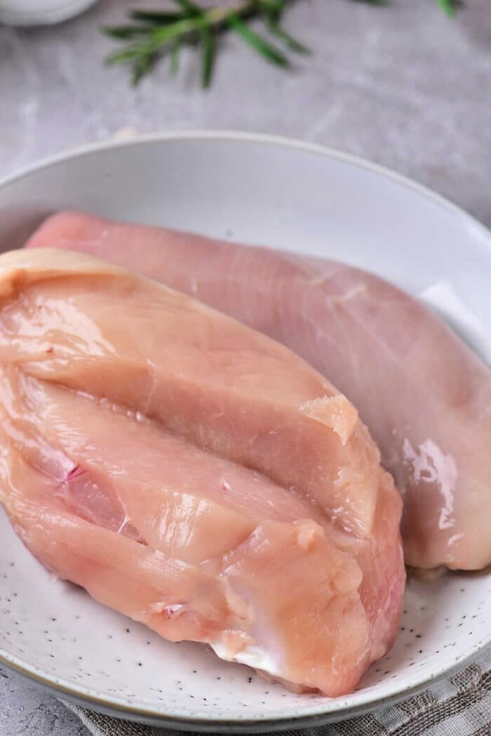 A white plate containing two raw chicken breasts set on a gray surface with green herbs in the background. One chicken breast is slightly pinker in color, while the other appears less so. The chicken breasts have slight marbling and visible fat, ready to be transformed into Crockpot Chicken with Italian Dressing.