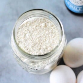 A jar filled with pancake mix sits on a surface, surrounded by eggs, as if preparing for something delicious, like Cracker Barrel Pancake Mix. A bottle is partially visible in the background.