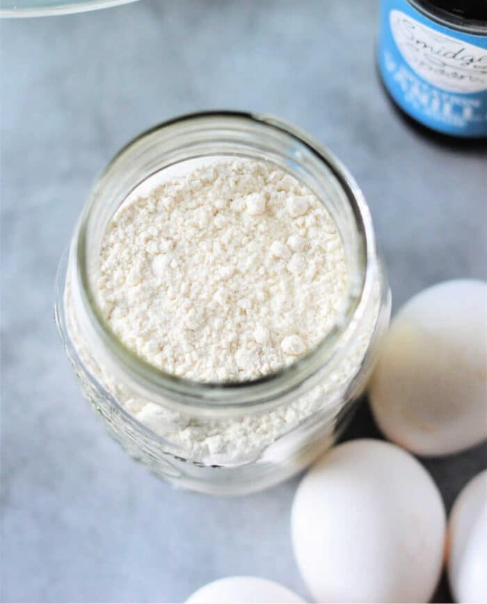 A jar filled with pancake mix sits on a surface, surrounded by eggs, as if preparing for something delicious, like Cracker Barrel Pancake Mix. A bottle is partially visible in the background.