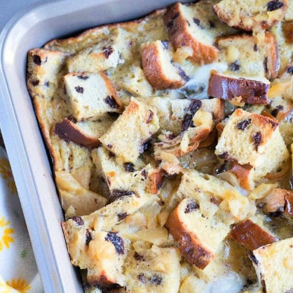 A baking pan filled with a delightful raisin bread pudding, featuring cubed bread pieces, some toasted. The mix of bread and creamy custard is studded with raisins and perhaps chocolate chips. The pan rests on a vibrant, colorful tablecloth.