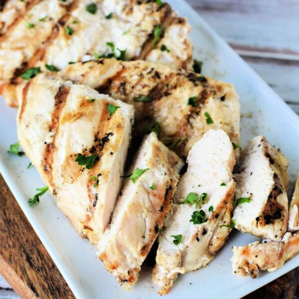 A plate of grilled chicken breast slices garnished with chopped parsley. The chicken, originally cooked as frozen chicken breast in an air fryer, has visible grill marks and the slices are arranged neatly on a rectangular white plate. The background includes a wooden surface.