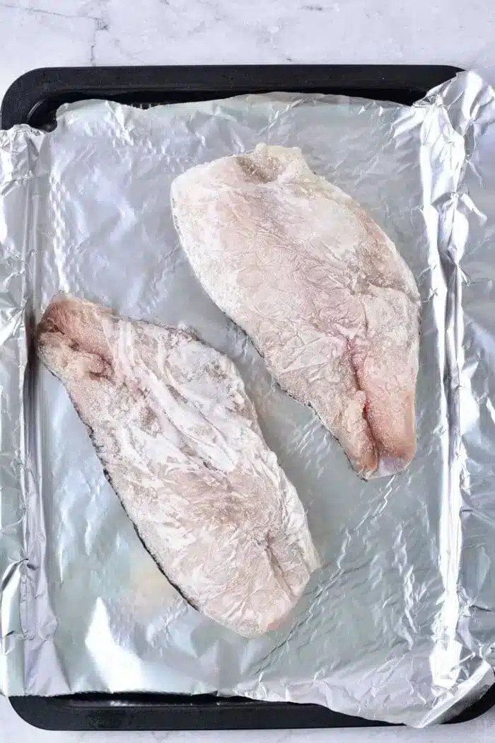 Two raw fish fillets rest on a baking sheet lined with aluminum foil, ready for baking. Their light, frosty appearance suggests they're frozen. The simple, light background enhances their fresh allure.