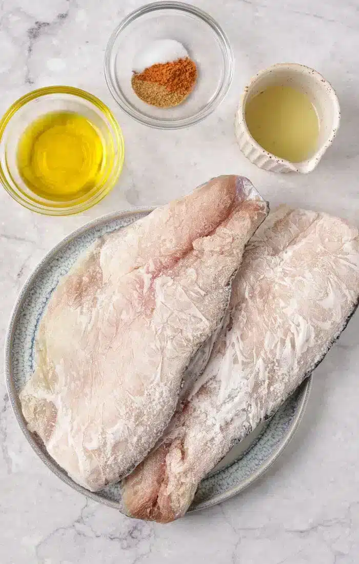 Two raw fish fillets sit on a plate, surrounded by small bowls containing olive oil, seasoning mix, and a light-colored liquid on a marble surface—ideal ingredients when baking frozen fish to perfection.