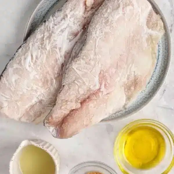 Two raw fish fillets on a plate are set on a marble surface, with small bowls of oil, lemon juice, and seasonings nearby—perfect preparation before baking frozen fish to perfection.