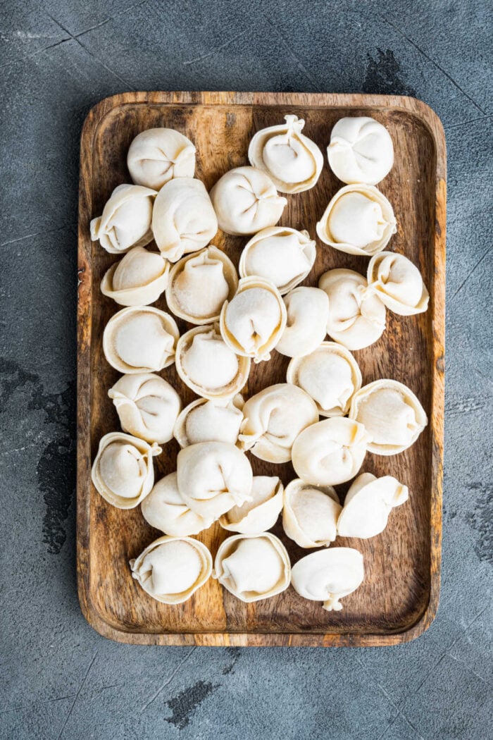 A wooden tray filled with uncooked dumplings on a textured gray surface, perfect inspiration for those exploring Crockpot Recipes with Tortellini.